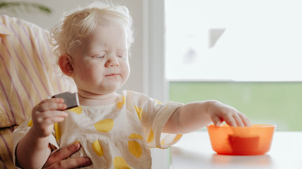 Cute toddler with light hair eating snacks from a bowl indoors. Bright natural light.